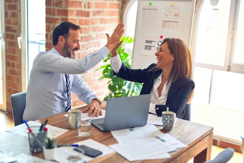 Two colleagues sharing a high five at a desk with a laptop